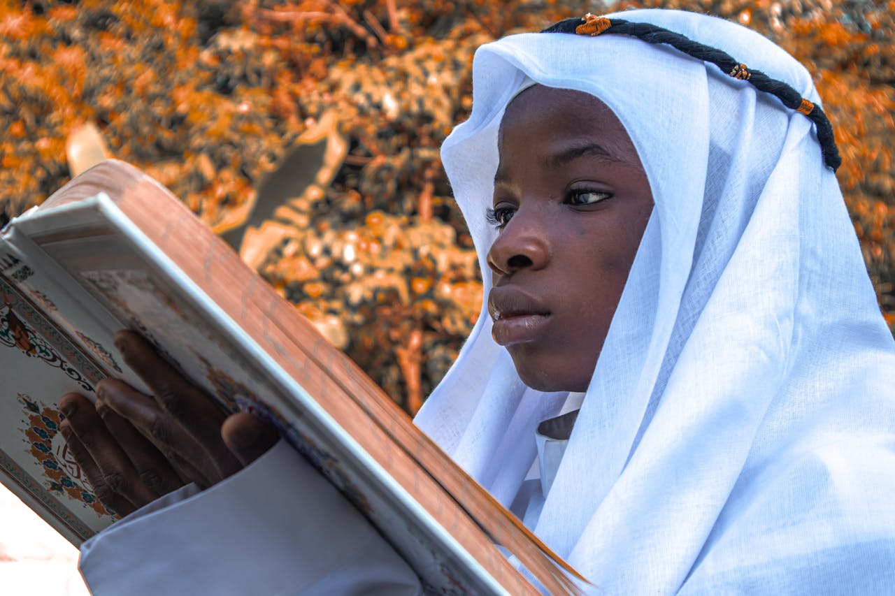 A young person in a traditional robe reading the Quran outdoors during autumn.